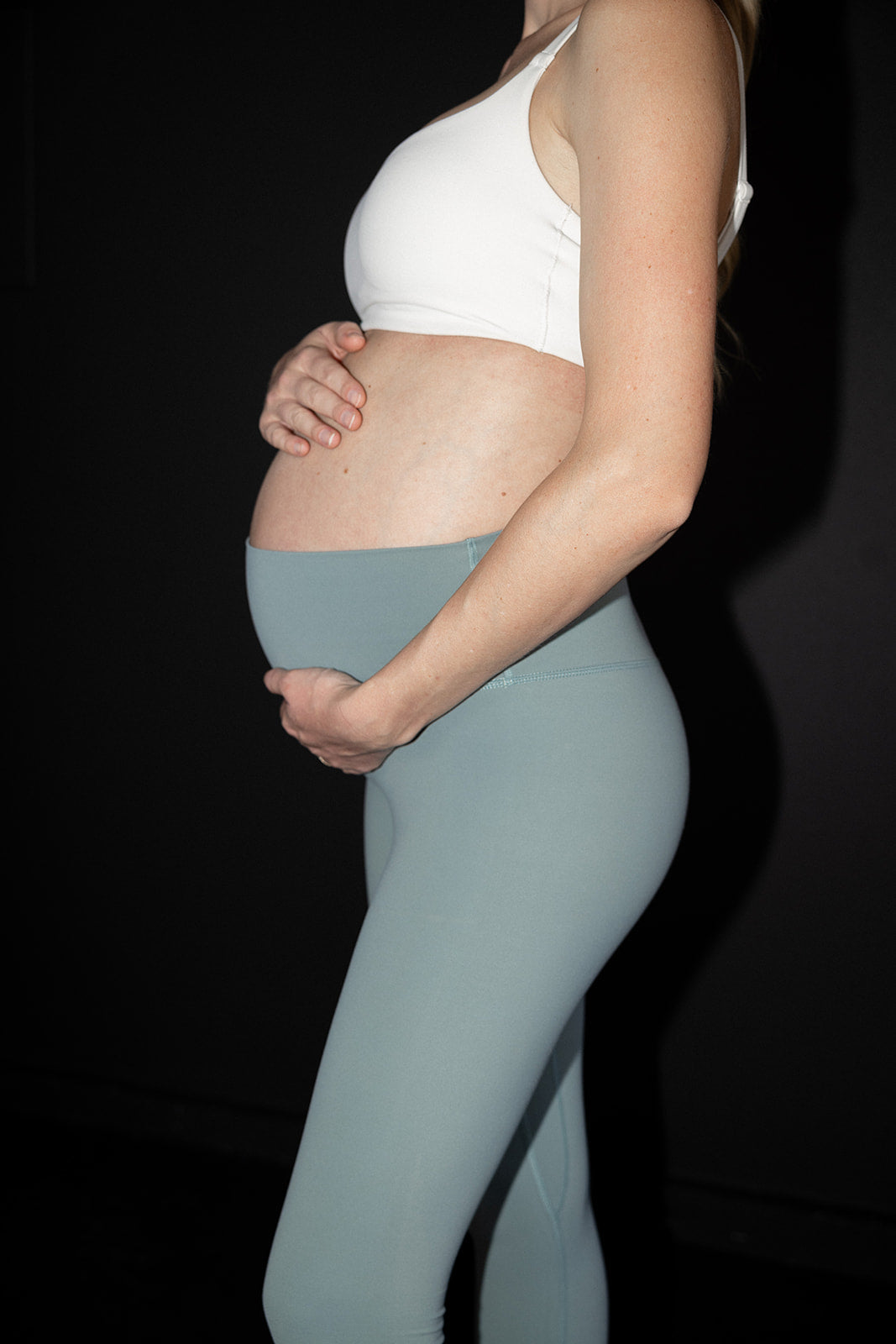 Momday image of pregnant womens body from the side in pregnant yoga pants and sports bra, holding the belly with both arms. This is for pregnancy workouts in third trimester. 